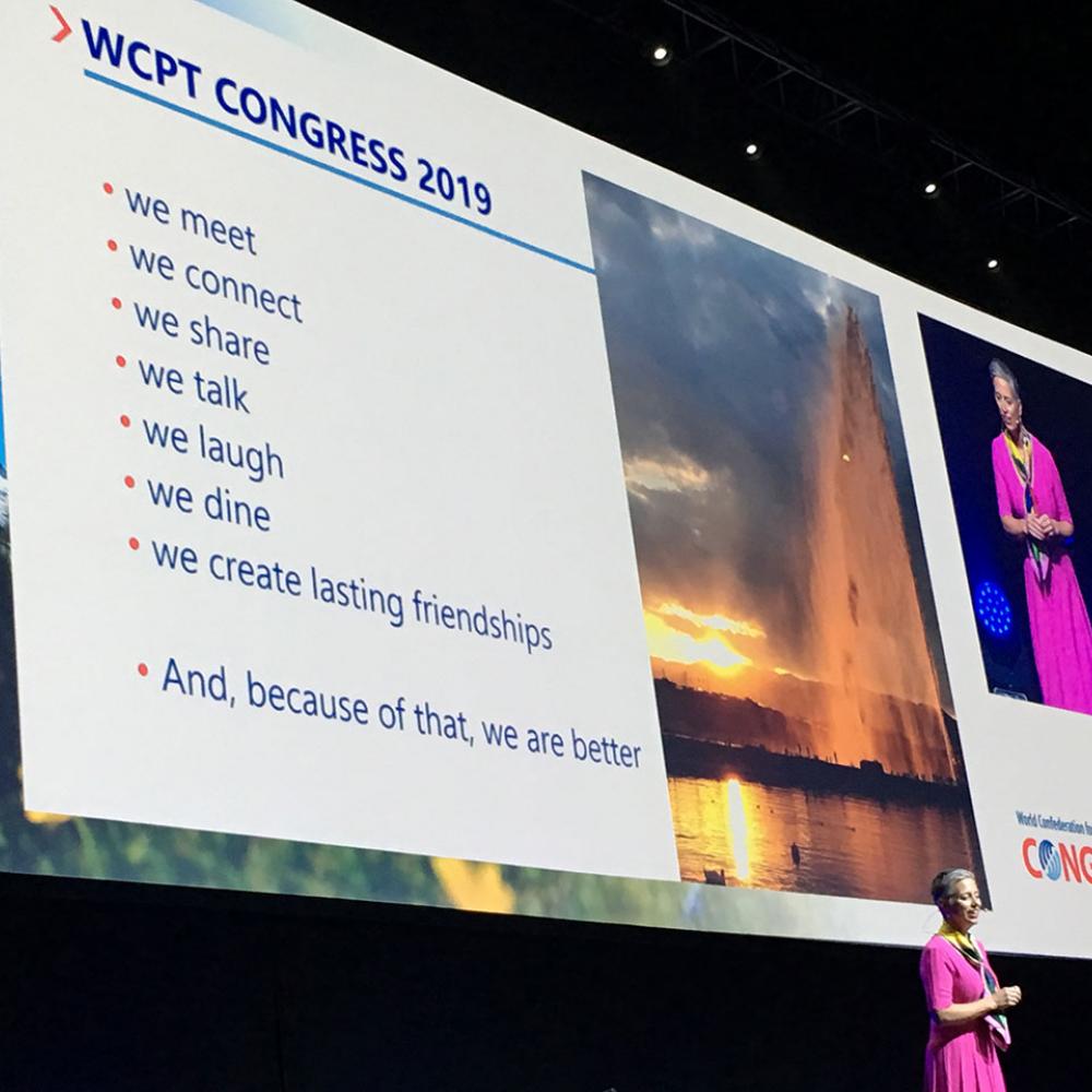 Emma Stokes on stage at Congress 2019, behind her a slide which says: WCPT Congress 2019, We meet, we connect, we share, we talk, we laugh, we dine, we create lasting friendships, and because of that we are better
