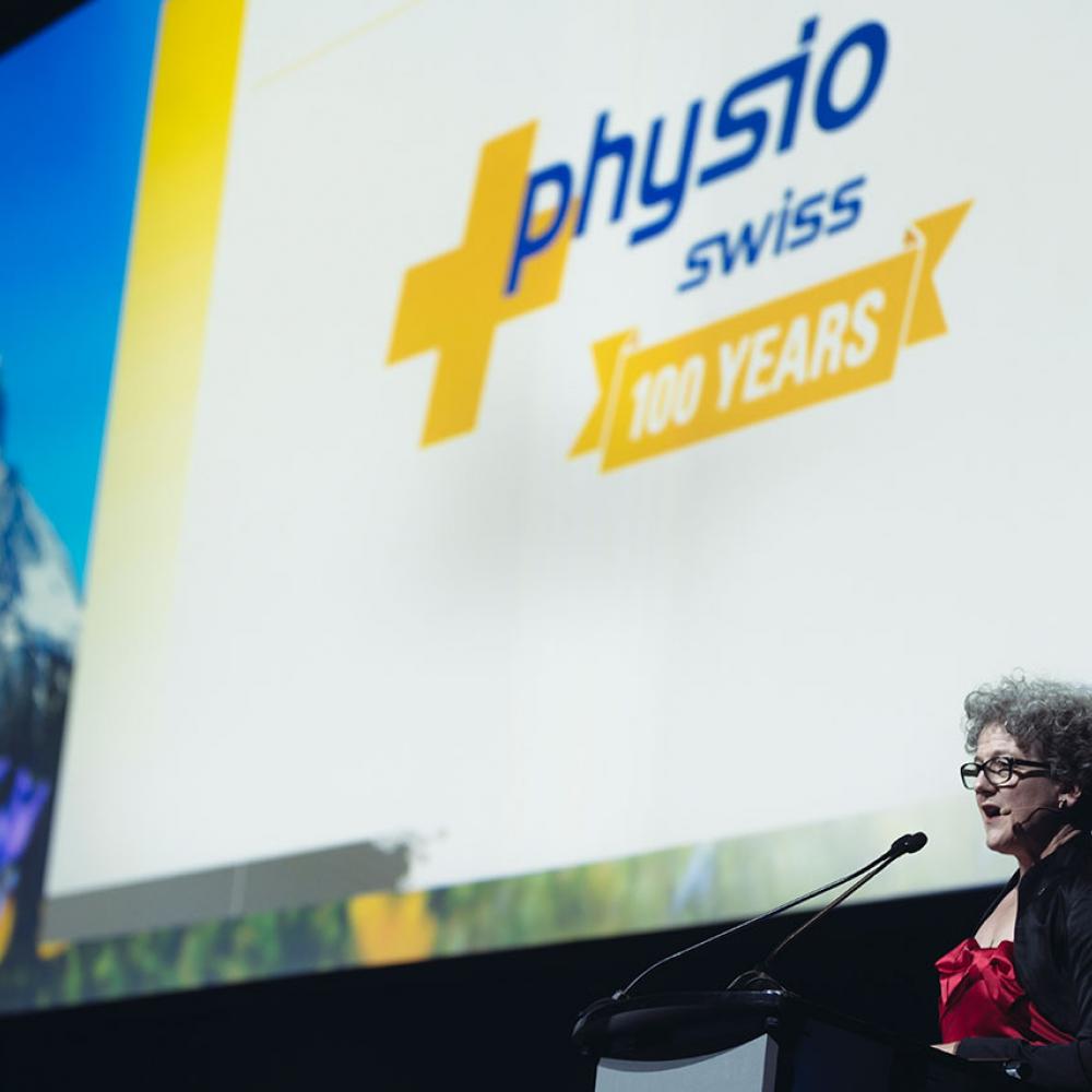 Seorang wanita berdiri di atas panggung berbicara ke mikrofon, di belakangnya ada slide yang bertuliskan: Physio Swiss, 100 years