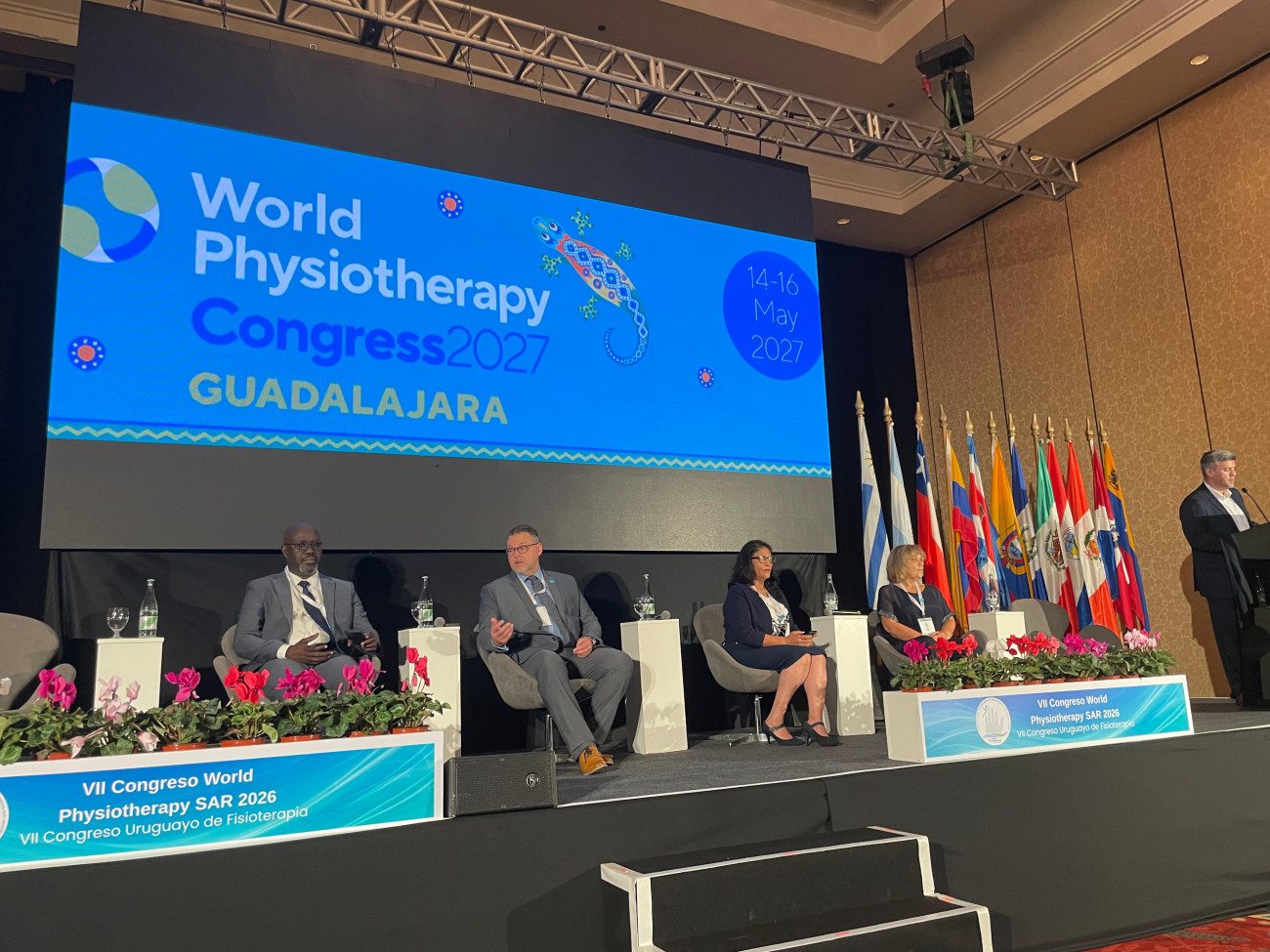 A group of panellists on stage discussing in front of a number of South American flags. The screen reads World Physiotherapy Guadalahara 2027
