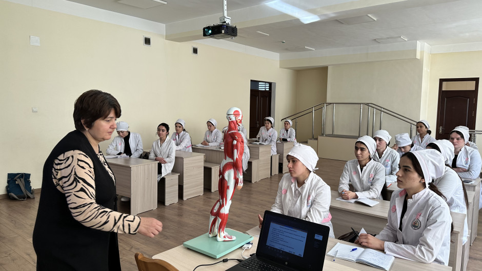 A physiotherapy instructor stands at the front of a classroom demonstrating a human muscle anatomy model while students in white medical uniforms sit at desks listening and taking notes.