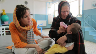 Two women sit on the floor, one bottle-feeds a baby